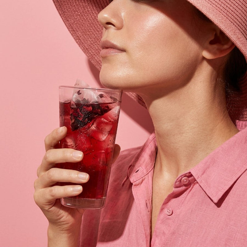 Close-up of glowing skin on a woman in a pink sun hat, holding a tall glass of iced hibiscus tea near her jawline
